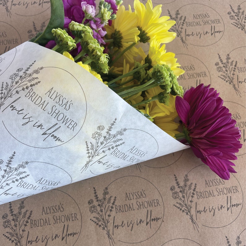 May include: A bouquet of flowers wrapped in white tissue paper with a floral design and the text "Alyssa's Bridal Shower Love is in Bloom".