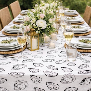 May include: A white tablecloth featuring a repeating black line-art pattern of oyster shells. The table is set with white plates on woven chargers, clear wine glasses filled with white wine, water glasses, and gold-framed lanterns with candles.