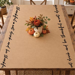 May include: A tan rectangular tablecloth featuring the black text "thankful eat drink and be thankful" printed along both long edges. A white ceramic pitcher filled with autumn flowers and three small pumpkins sits in the center.