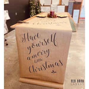 May include: A brown paper table runner with the text "Have yourself a merry little Christmas" in elegant script. The runner is draped over a wooden table, with a small candle and berries on top. The Red Barn Canvas logo is in the bottom right corner.