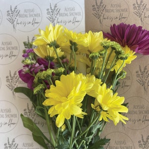 May include: A bouquet of yellow and purple flowers wrapped in brown paper with circular stickers that say "Alyssa's Bridal Shower Love is in Bloom".