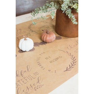 May include: A brown paper table runner with handwritten text that says "Thankful, Grateful, Blessed" and "Thankful for my husband". The runner is decorated with small white and brown pumpkins and leafy sprigs.