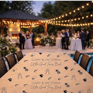 May include: A tan tablecloth with black illustrations of champagne glasses, lemons, and other party-themed graphics. The text "CORY & VICTORIA" and "Let the Good Times Ball" are printed on the cloth. The table is set for an outdoor event.