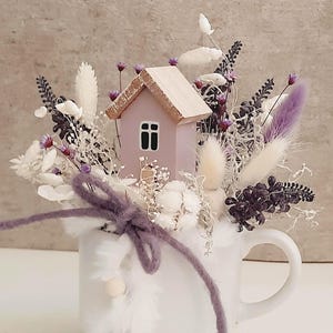 Op de afbeelding: Een witte mok gevuld met een bloemstuk met een klein roze huis, droogbloemen en paarse accenten. Het arrangement bevat witte, paarse en crèmekleurige elementen, met een paarse vilten strik rond de mok.