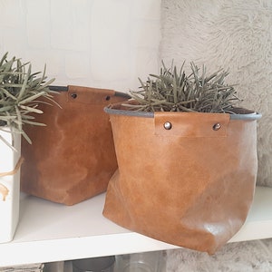 Op de afbeelding: Twee bruine leren plantenpotten met grijze afwerking en metalen klinknagels. De potten staan op een wit rek met een wit, pluche tapijt op de achtergrond.