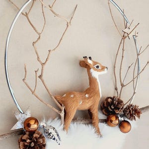 Könnte beinhalten: Ein dekorativer Weihnachtskranz mit einer braunen Hirschfigur mit weißen Punkten, vor einem Hintergrund aus Zweigen und einem silbernen Reifen. Der Kranz enthält Tannenzapfen, Ornamente und einen Stern.