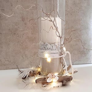 Könnte beinhalten: Eine dekorative Glasvase gefüllt mit Sand, weißen Steinen und einer großen weißen Kerze. Die Vase ist mit Seil umwickelt und mit glitzernden Zweigen und Lichterketten verziert. Muscheln, Federn und Treibholz vervollständigen das Küsten-Themen-Arrangement.