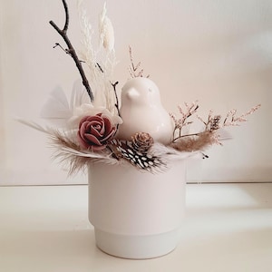 Op de afbeelding: Een witte keramische vogelbeeldje staat op een witte vaas, omringd door gedroogde bloemen, veren en een roos. De opstelling bevat een kleine dennenappel en een tak, waardoor een natuurlijke, decoratieve presentatie ontstaat. De vaas is cilindrisch met een getrapte basis.