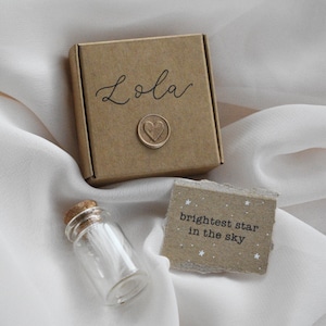 May include: A brown gift box with the name "Lola" in cursive and a heart seal. A small glass bottle with a cork stopper and a tag that reads "brightest star in the sky" are also present. The items are on a white fabric.