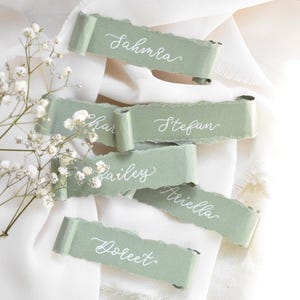 May include: Sage green place cards with torn edges, each bearing a guest's name in elegant white calligraphy. The cards are arranged on a white surface, with delicate white flowers adding a touch of nature.