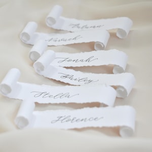 May include: White paper scrolls with torn edges, each bearing a name in elegant calligraphy. Names include Autumn, Hannah, Jonah, Bailey, Stella, and Florence. The scrolls are arranged on a soft, off-white fabric, creating a delicate and romantic aesthetic.
