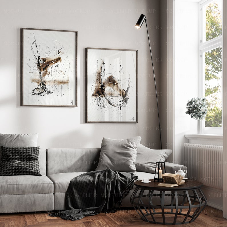 May include: A living room interior with a gray sofa, two black and white abstract paintings, a black floor lamp, a black coffee table, a throw blanket, and a window with a view of trees.