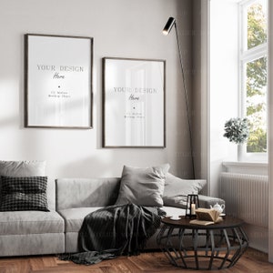 May include: Two framed posters with the text "YOUR DESIGN Here" and "C2 Atelier Mockup Store" hanging on a white wall above a gray couch with pillows and a throw blanket. A black floor lamp with a round shade is in the corner of the room. A black metal coffee table with a book and a teapot on top is in front of the couch.