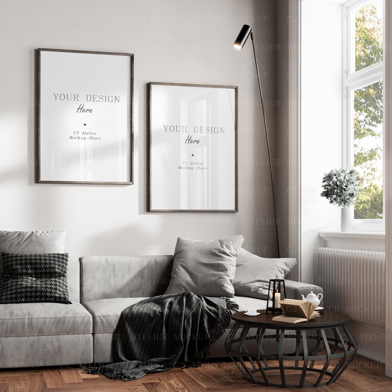 May include: Two framed posters with the text "YOUR DESIGN Here" and "C2 Atelier Mockup Store" hanging on a white wall above a gray couch with pillows and a throw blanket. A black floor lamp with a round shade is in the corner of the room. A black metal coffee table with a book and a teapot on top is in front of the couch.