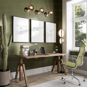 Op de afbeelding: Een thuiskantoorwerkplek met een houten bureau, een groene cactusplant, drie ingelijste posters met de tekst "Your Design Here", een laptop, een koptelefoon, een glas water, een stapel boeken, een metalen lamp met een ronde kap en een groene bureaustoel.