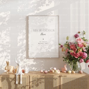 Op de afbeelding: Een witte frame mockup met de tekst "YOUR DESIGN Here" en "C2 Atelier Mockup Store" op een witte muur met een boeket roze bloemen in een glazen vaas en een houten kast met witte keramische konijnenbeeldjes en pastelkleurige paaseieren.