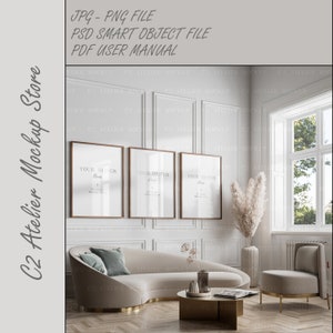 May include: Three framed posters with the text "YOUR DESIGN Here" hanging on a white wall above a curved white sofa with a blue pillow. A tall vase with dried pampas grass is next to the sofa. A small round gold table with a book and a glass vase is in front of the sofa. A white armchair with gold legs is in the corner of the room. A window with a view of trees is in the background.