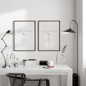 Puede incluir: Dos carteles enmarcados en marrón con el texto "YOUR DESIGN Here C2 Atelier Mockup Store" colgados en una pared blanca sobre un escritorio blanco con una silla negra, una lámpara, un jarrón con una pluma, una taza de café y un plato de bayas rojas.