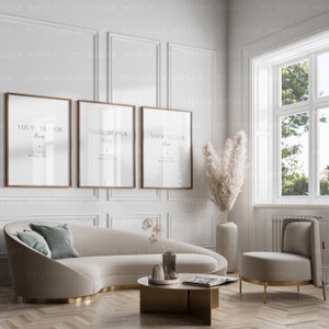 May include: Three framed posters with the text "Your Design Here" hanging on a white wall with decorative molding. A white curved sofa with a teal blue pillow sits in front of the posters. A small round gold table with a magazine and a vase with dried flowers sits in front of the sofa. A white chair with gold legs sits to the right of the sofa. A large vase with dried pampas grass sits in the corner of the room.