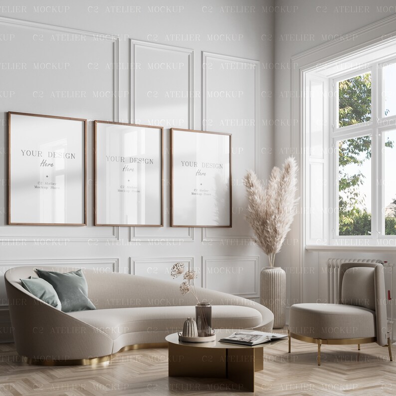May include: Three framed posters with the text "Your Design Here" hanging on a white wall with decorative molding. A white curved sofa with a teal blue pillow sits in front of the posters. A small round gold table with a magazine and a vase with dried flowers sits in front of the sofa. A white chair with gold legs sits to the right of the sofa. A large vase with dried pampas grass sits in the corner of the room.