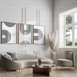 May include: Three framed black and white abstract prints with geometric shapes hang on a white wall above a curved beige sofa with a light blue throw pillow. A vase with dried pampas grass and a small coffee table with a book and a decorative bowl are in the room.