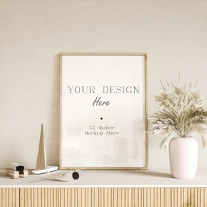 May include: A gold framed picture mockup with a white background and the text "YOUR DESIGN Here" and "C2 Atelier Mockup Store". The frame is on a light wood shelf with a white surface. A small wooden sailboat and a toy airplane are on the shelf. A vase with dried pampas grass is on the right side of the shelf.
