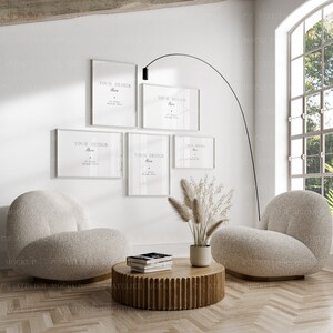 Könnte beinhalten: Fünf weiße Bilderrahmen mit dem Text "Your Design Here" hängen an einer weißen Wand. Die Rahmen sind in einer rechteckigen Form angeordnet. Im Vordergrund stehen zwei weiße Sessel und ein Holz-Couchtisch mit Büchern darauf. Der Raum hat ein großes Fenster mit Blick auf Bäume.