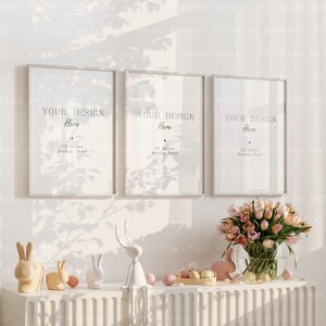 以下が含まれることがあります： 白い壁に、「Your Design Here」と「C2 Atelier Mockup Store」の文字が書かれた白い写真フレームが3つ掛かっています。フレームは窓からの影に囲まれています。フレームの下には、ピンクのチューリップの花瓶、パステルカラーのイースターエッグのボウル、白いウサギの置物が3つあります。
