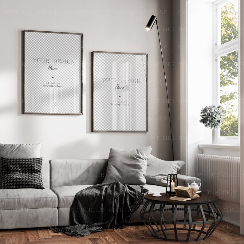 May include: Two framed posters with a transparent background, the text "YOUR DESIGN Here" is visible on each poster. The posters are hanging on a white wall above a gray couch with black and gray throw pillows and a black throw blanket. A black floor lamp with a cylindrical shade is in the corner of the room. A black metal coffee table with a geometric design is in front of the couch.