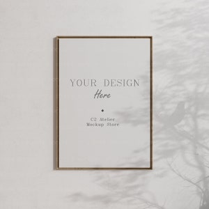 Puede incluir: Una maqueta de póster enmarcada en dorado con el texto "YOUR DESIGN Here" y "C2 Atelier Mockup Store" en una pared blanca con la sombra de una rama de árbol.