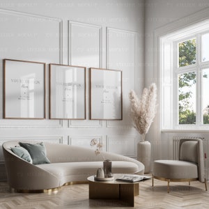 May include: Three framed pictures with the text "Your Design Here" hanging on a white wall above a curved beige sofa with a teal blue pillow. A vase with dried pampas grass is on the right side of the image.