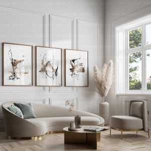 May include: Three abstract black and white framed paintings with brown accents hang on a white wall above a curved beige sofa with a teal blue throw pillow. A vase with dried pampas grass sits on the floor next to a beige armchair.