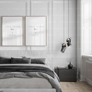 Op de afbeelding: Twee ingelijste posters met de tekst "YOUR DESIGN Here" en "C2 Atelier Mockup Store" hangen aan een witte muur boven een grijs bed met zwarte kussens en een grijs dekbed. Een zwarte bijzettafel met een vaas met gedroogde takken staat naast het bed. Twee zwarte hanglampen met gouden accenten hangen aan het plafond.