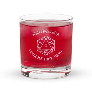 May include: A clear glass tumbler filled with a red liquid, possibly a cocktail. The glass features white text that reads "Yeah I rolled a 1, pour me that drink" around a white D20 die graphic. The glass is set against a white background.