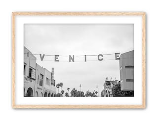 May include: Black and white photograph of a street scene in Venice, California. The word "VENICE" is spelled out in large letters strung across the street. Palm trees line the street.