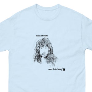 May include: Light blue t-shirt with a black and white illustration of a man with long hair. The text "music and dreams" and "robert lester folsom" are printed on the shirt.