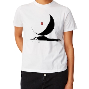 May include: White t-shirt featuring a black crescent moon design with the number 6 in red. Below the moon is a black silhouette of a person lying down. The shirt is a crew neck style.