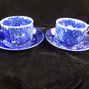 May include: Two blue and white teacups and saucers with a mottled glaze. The cups and saucers are on a black surface.