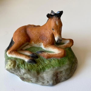 Puede incluir: Figura de cerámica de un potro en reposo. El potro es marrón con crin y pezuñas negras, tumbado sobre una base verde y gris. La figura es detallada y parece pintada a mano, mostrando una representación realista del animal.