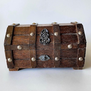 Puede incluir: Un cofre de madera rectangular con acabado marrón oscuro, que presenta una tapa curva y listones verticales. El cofre está decorado con tachuelas metálicas de tono plateado e incluye un cierre central de metal ornamentado y un emblema.