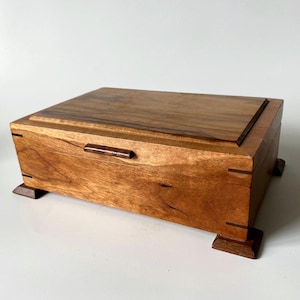 May include: A rectangular wooden box with a hinged lid and a small horizontal handle. The box features a light brown wood grain with darker wood accents on the corners, edges, and four small feet. The lid has a raised centre panel.