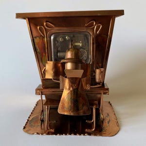 May include: A copper-toned metal music box featuring a stylized figure seated at a piano. The figure wears a hat and sits on a bench. The piano has a visible internal mechanical movement behind a clear window, with decorative accents on the base.