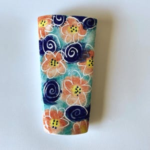May include: A rectangular ceramic vase featuring a hand-painted pattern of orange flowers with yellow centers and dark blue spiral accents against a textured teal and light blue background. The vase has a flat top edge and a slightly tapered base.