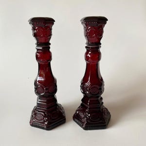 May include: A pair of deep red glass candle holders featuring a pressed pattern with circular and diamond-shaped motifs. Each candlestick has a hexagonal base, a tapered stem, and a wide top rim, all crafted from translucent dark red glass.