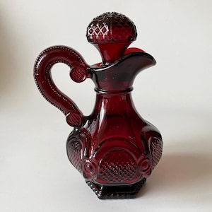 Pode incluir: Uma jarra de vidro vermelho profundo com um padrão de diamante texturizado no corpo e uma tampa a condizer. O recipiente tem uma pega curva com detalhes circulares em relevo e um bico largo, com um acabamento em tom rubi translúcido.