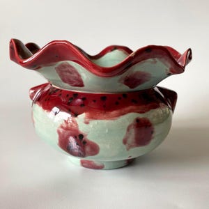 May include: A ceramic bowl with a ruffled rim, painted in shades of red and mint green. The bowl features a rounded base and a decorative, wavy edge. Red accents and dark speckles add detail to the design. A unique piece for home decor.