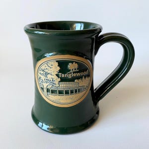 May include: A dark green ceramic mug with a curved handle. The mug features a gold oval design with the word "Tanglewood" and a landscape illustration. The mug is approximately 4 inches tall.