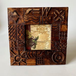 May include: A square wooden picture frame featuring a dark brown finish and intricate hand-carved geometric patterns across its border. The frame holds a print depicting a dark vase with green leaves and white flowers against a gold background.