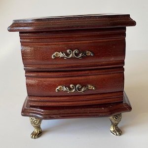 May include: A small, dark brown wooden chest with two drawers, each featuring an ornate, curved brass-toned metal handle. The chest is supported by four decorative, gold-toned metal feet shaped like animal paws, providing a raised base.