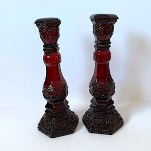 Puede incluir: Un par de candelabros de vidrio rojo oscuro con un patrón prensado de motivos circulares y geométricos. Cada candelabro tiene una base cuadrada, un tallo cónico y una parte superior cuadrada, con un acabado translúcido y detalles en relieve.
